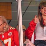 Taylor Swift in a white tank top and red jacket makes a thumbs up as she stands in a suite next to Donna Kelce in a Travis Kelce Chiefs jersey